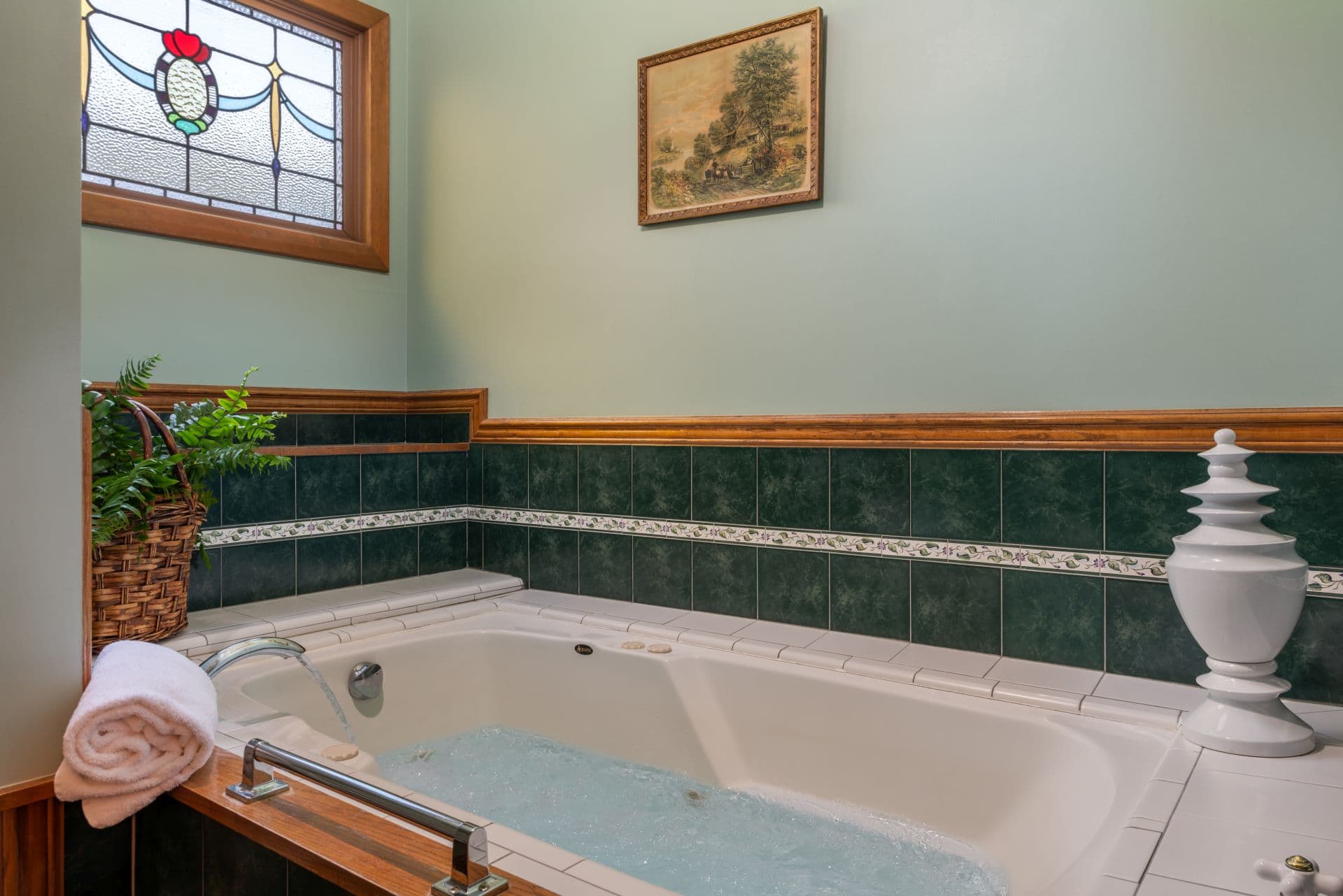 Bathroom with Jacuzzi soaking tub bordered by green and white subway tiles with a stained glass window above.