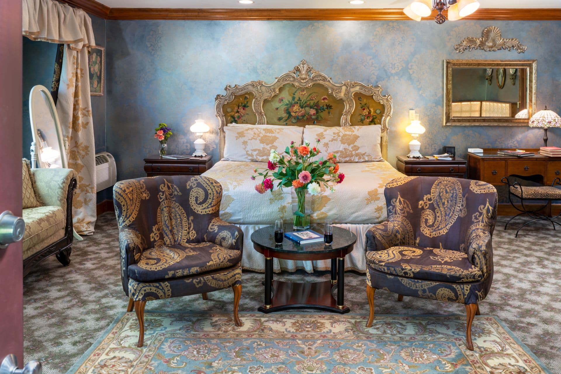 A pair of upholstered armchairs flanking a round table topped with a vase of flowers at the foot of an ornate king-size bed.