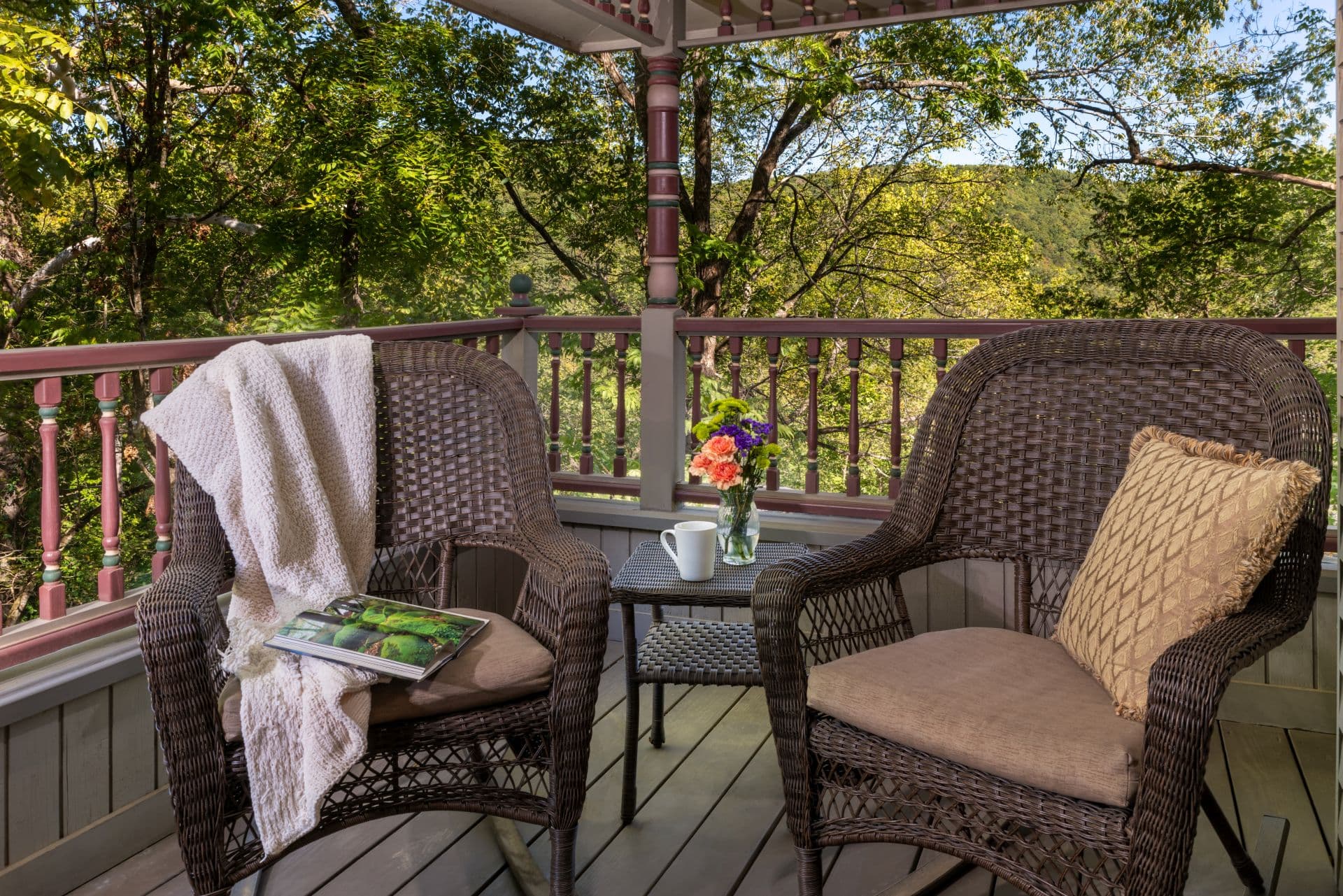 Two wicker armchairs and a small occasional table on a private balcony overlooking lush green trees.