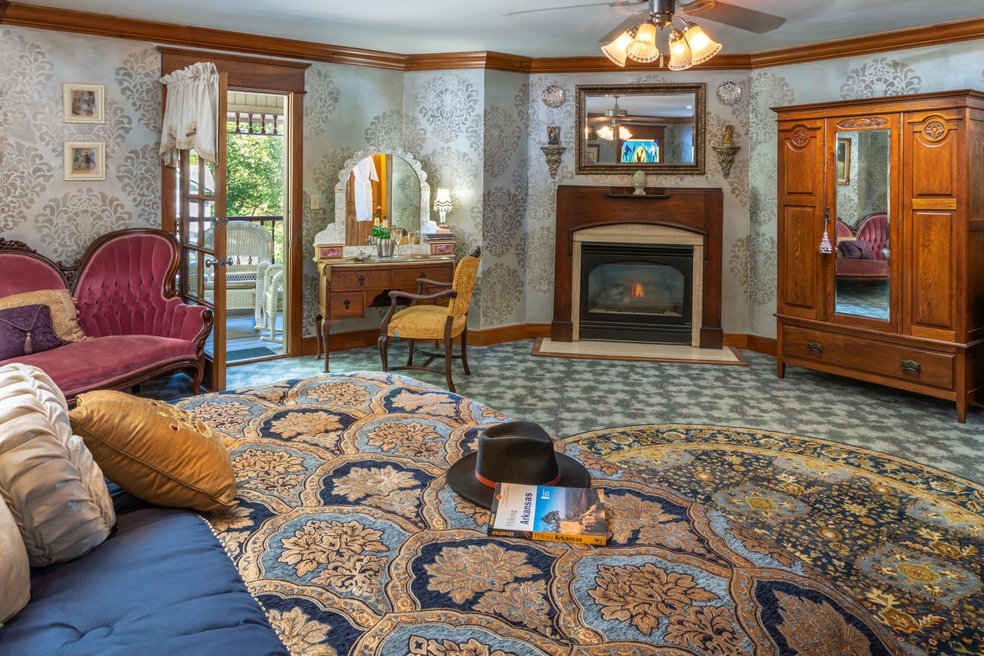 Luxurious bedroom with fireplace, antique armoire, antique velvet sofa, and vanity with mirror opposite decadent king-size bed.