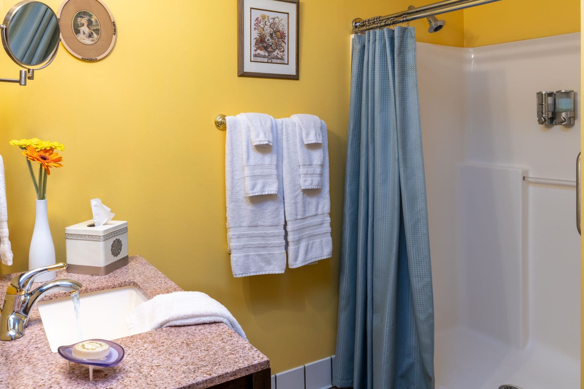 Bathroom with rich yellow walls, vanity with sink, vintage-style shaving mirror, and shower with plush white towels hanging nearby.