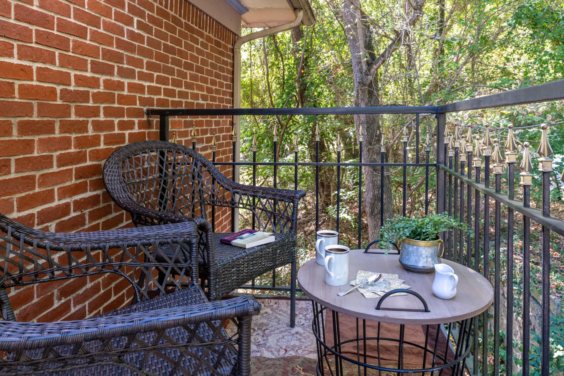 A private balcony with two wicker chairs and a round occasional table with views of trees and foliage.