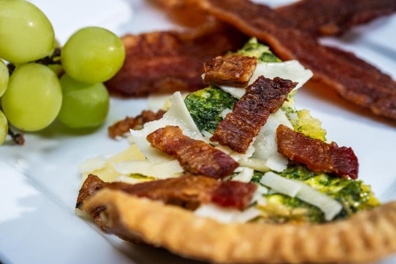 A plate featuring a slice of frittata topped with crispy bacon, alongside green grapes and more bacon strips.