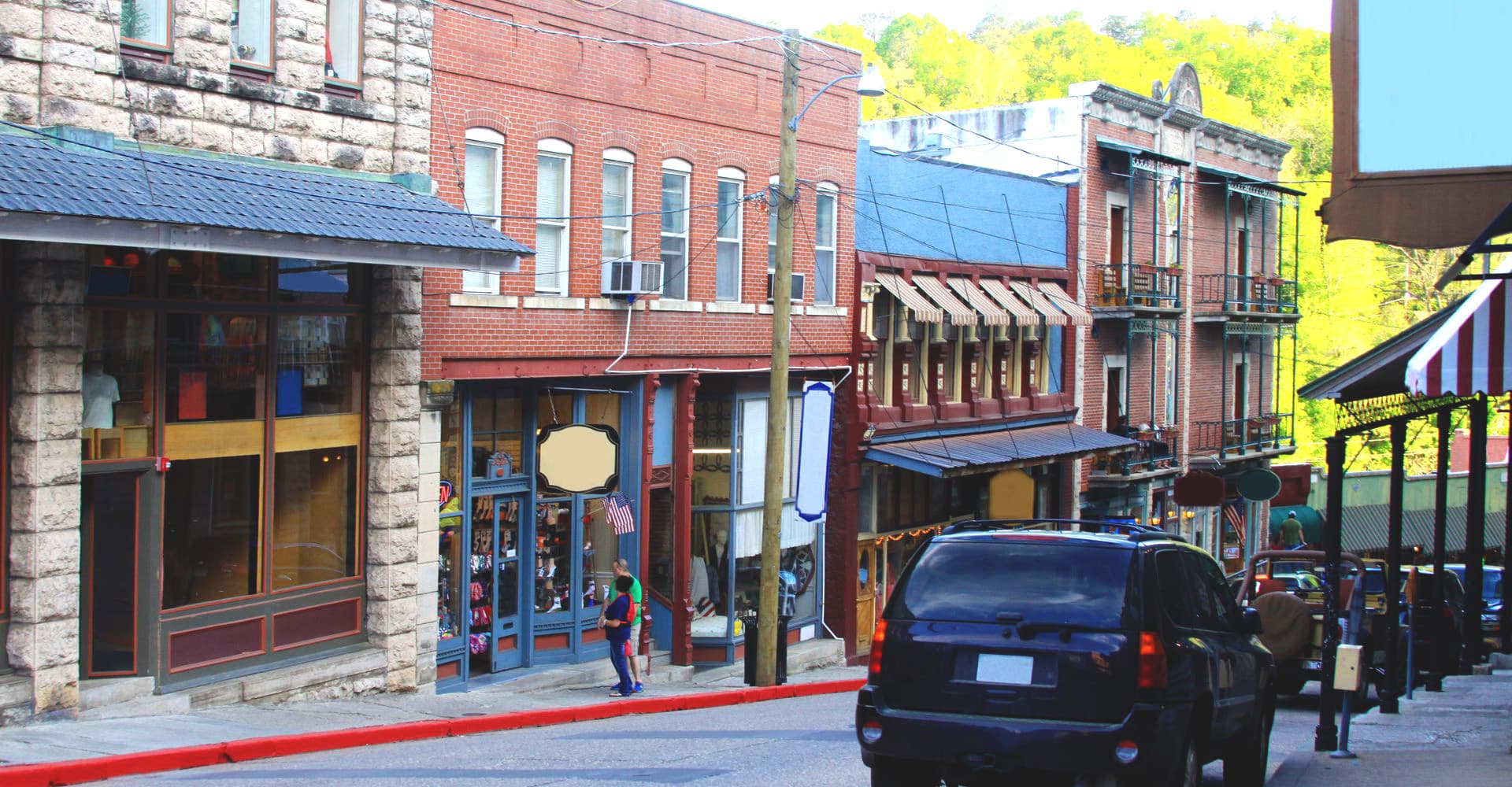 A street scene featuring historic buildings, shops, and parked cars on a sloped road.
