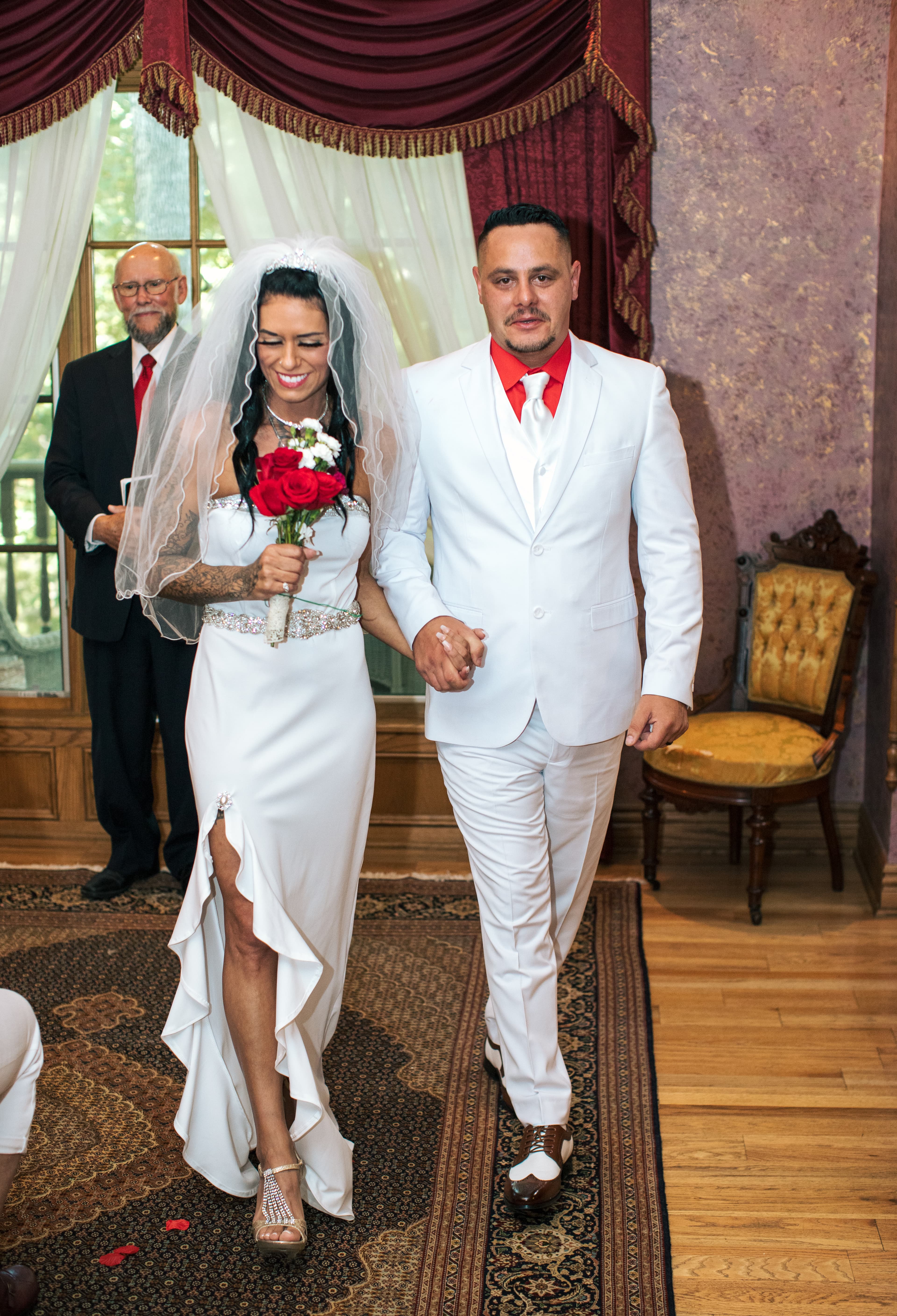 A smiling bride in a white dress and a groom in a white suit hold hands, walking together in a decorated venue.
