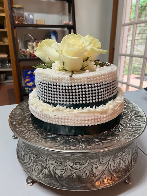 A decorative two-tier cake adorned with pearls, black ribbon, and a white rose, sitting on an ornate silver stand.