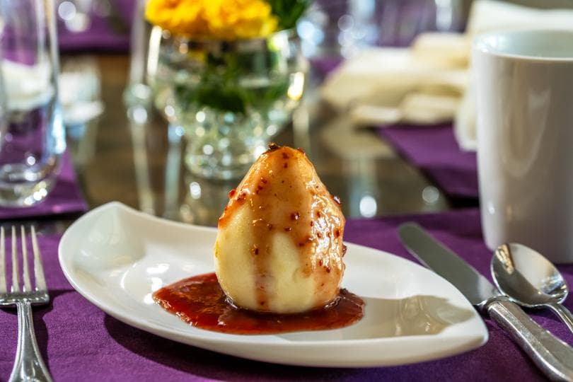 A poached pear drizzled with sauce sits on a white plate on a neatly set table.
