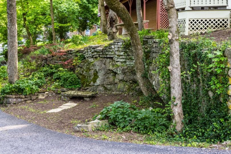 A stone bench sits among lush greenery and trees beside a gravel path.