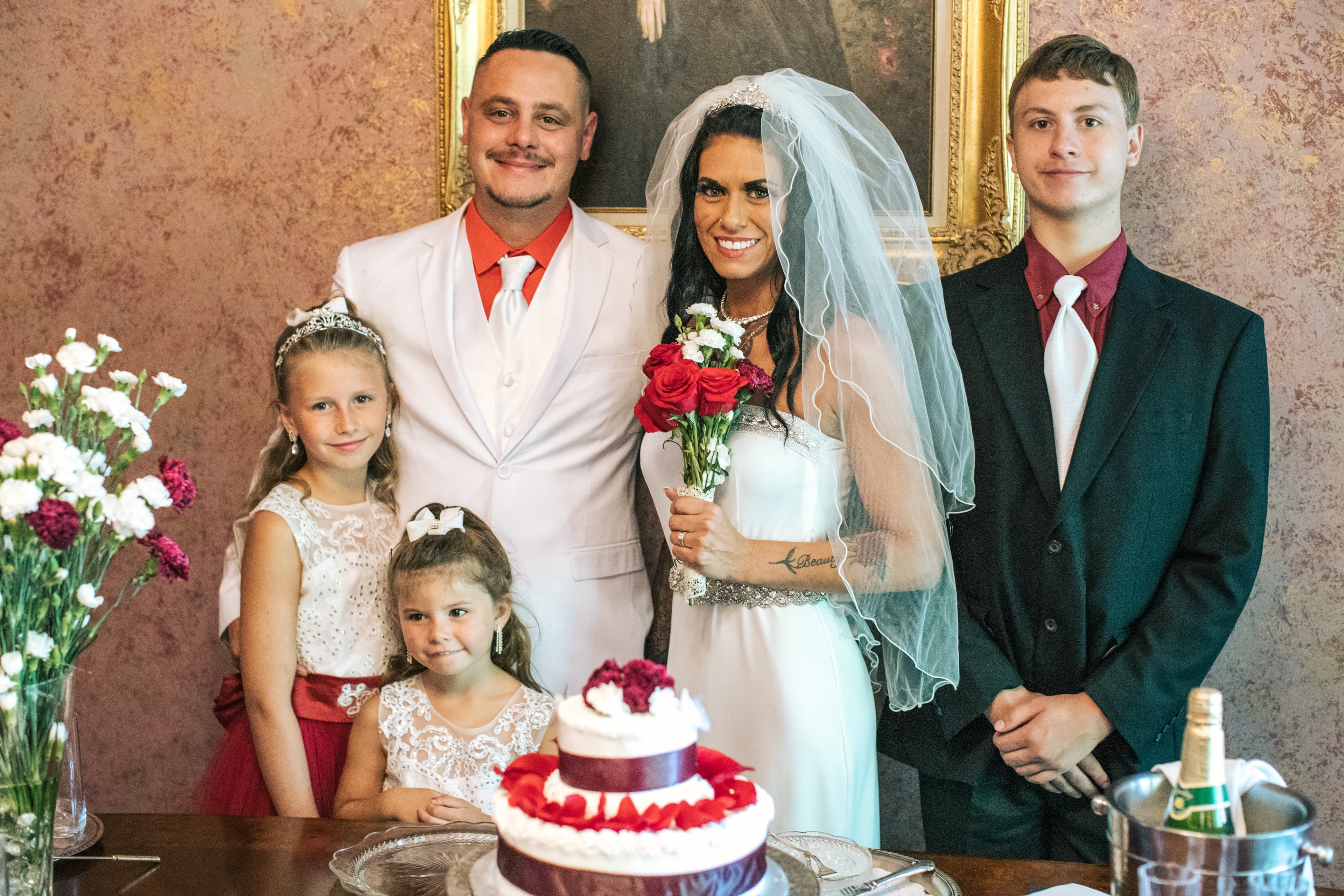 A bride and groom stand with two children and a young man beside a wedding cake.