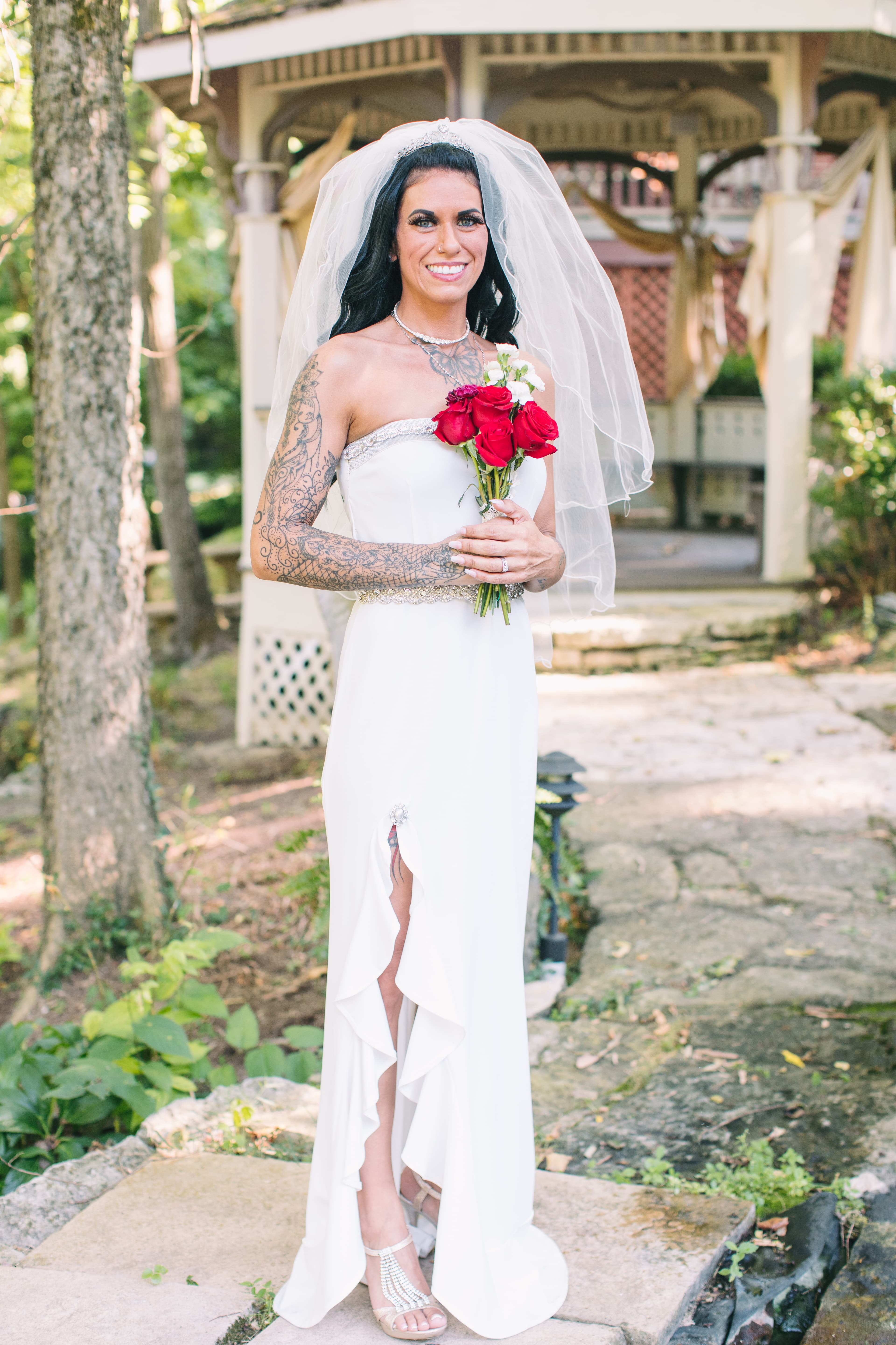 A smiling bride in a white gown holds a bouquet of red roses while standing near a gazebo in a lush outdoor setting.