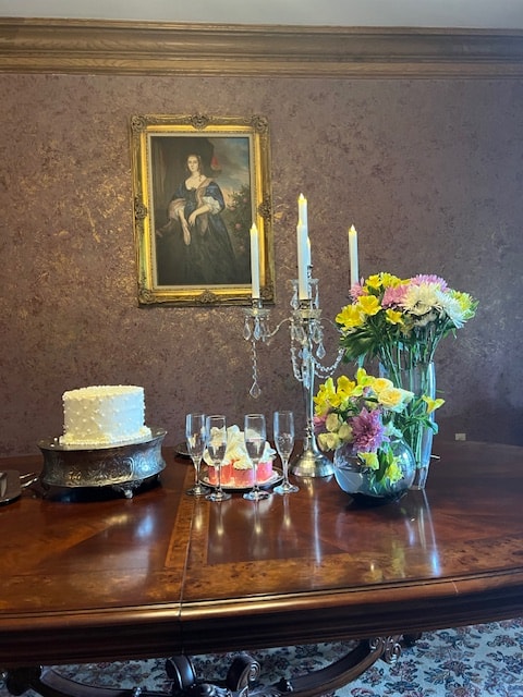 A beautifully set dining table with a white cake, flutes, a floral arrangement, and vintage candlesticks, against a backdrop of a framed painting.