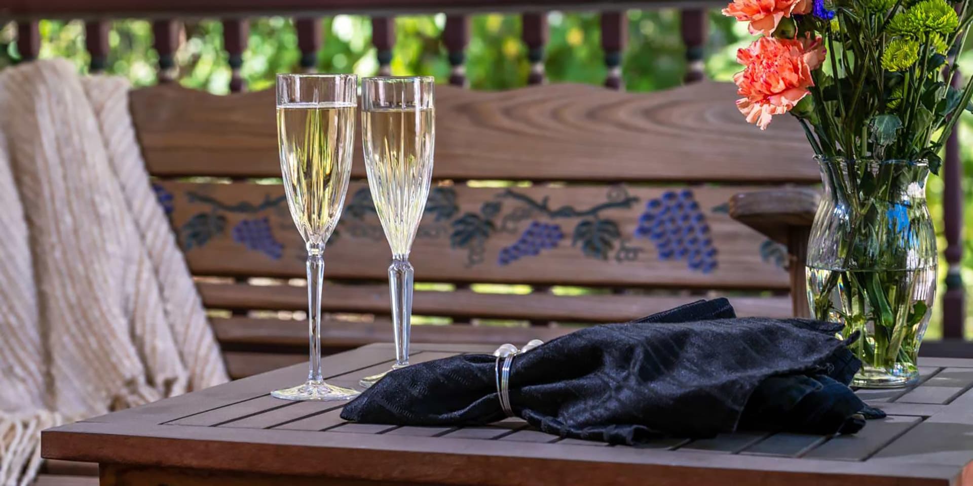Two glasses of sparkling wine sit on a wooden table beside a vase of flowers and a folded napkin.