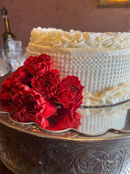 A white cake decorated with intricate frosting sits on a mirrored surface, accompanied by a bunch of vibrant red carnations.