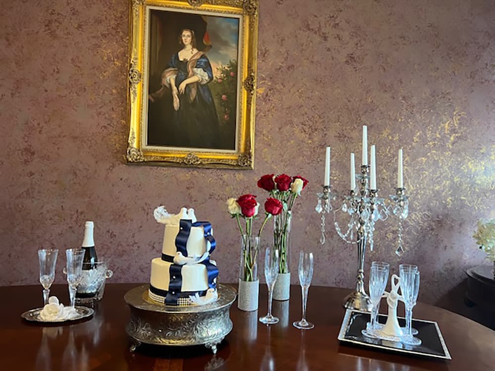 A decorated table featuring a wedding cake, champagne, roses, and elegant glassware, with a portrait in the background.