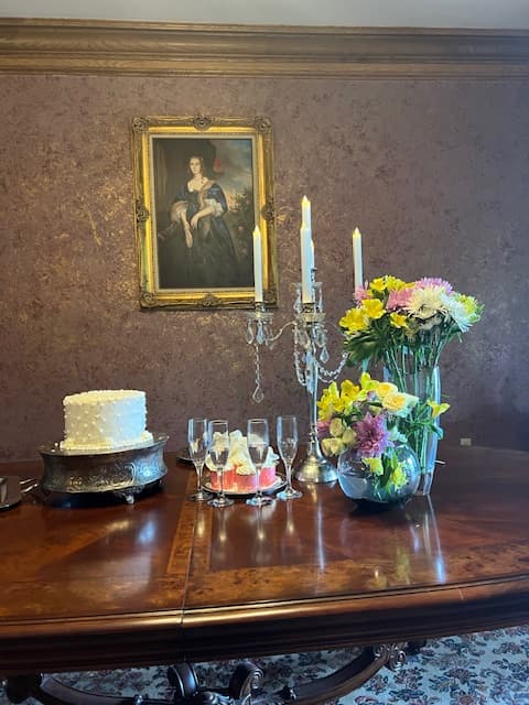 A table set for a celebration, featuring a decorated cake, candles, champagne glasses, and a vibrant bouquet, with a classic portrait in the background.