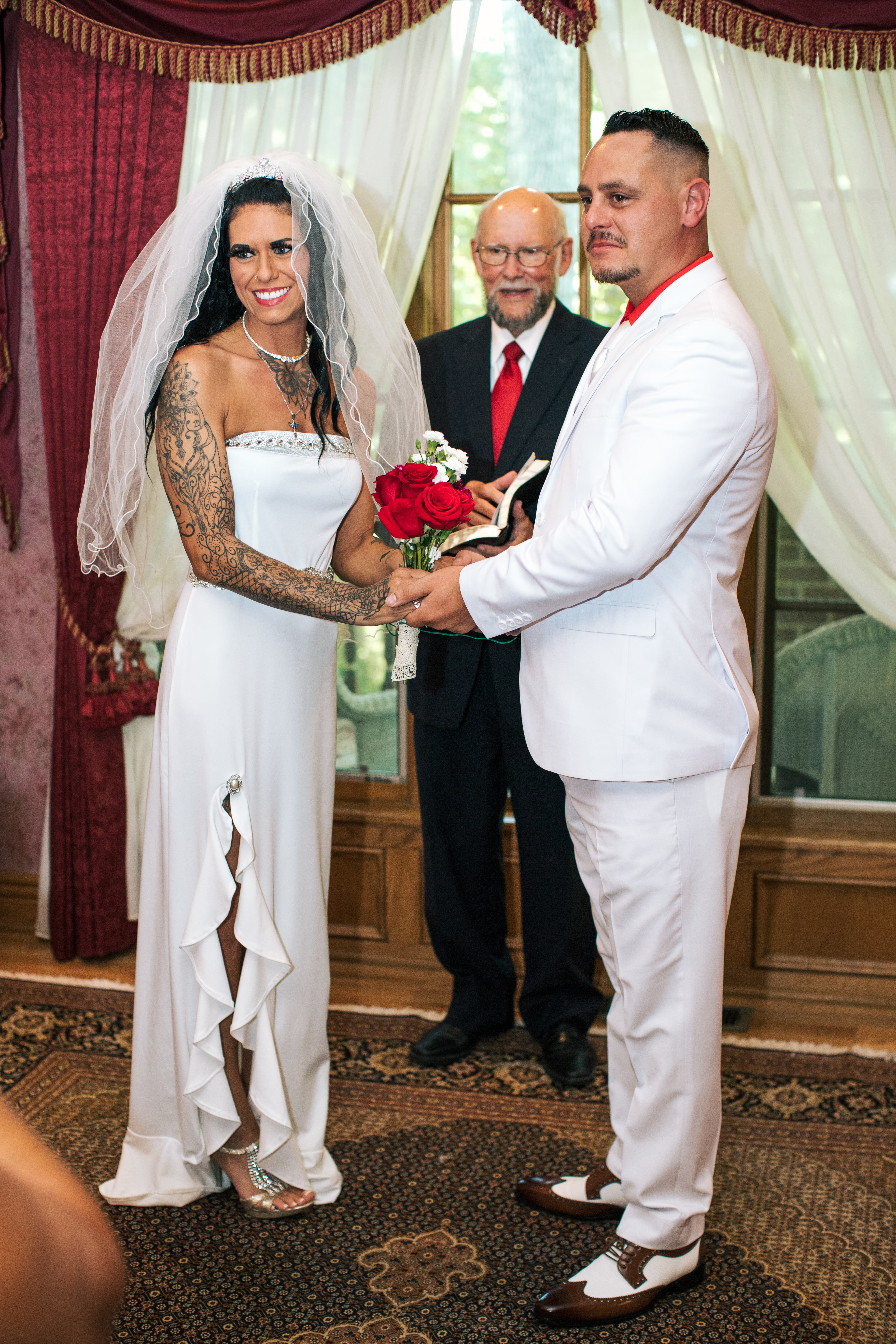 A bride in a white dress holds a bouquet while exchanging vows with a groom in a white suit, with an officiant in the background.