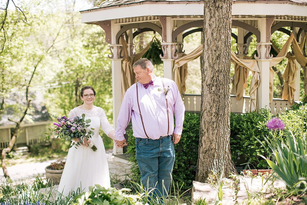 A bride in a white dress holds a bouquet while walking hand-in-hand with a man in a purple shirt near a gazebo.