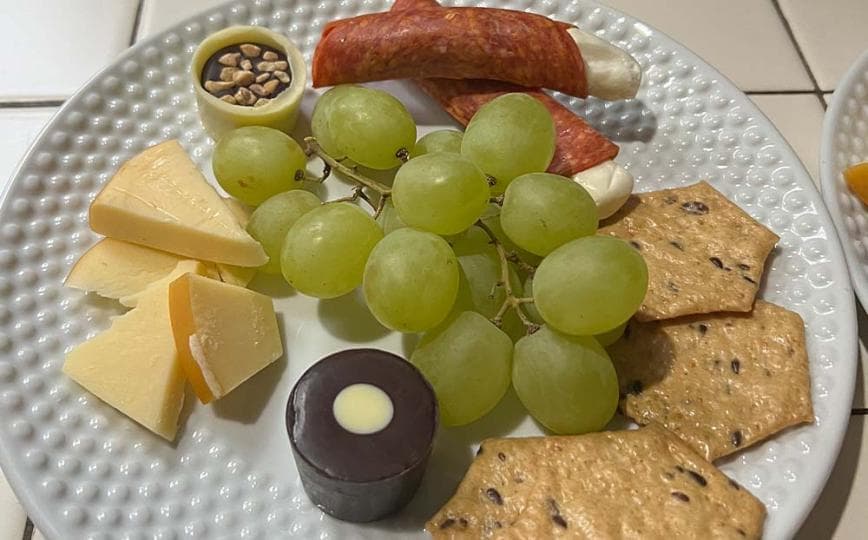 A plate featuring green grapes, assorted cheeses, salami, crackers, and a small chocolate treat.