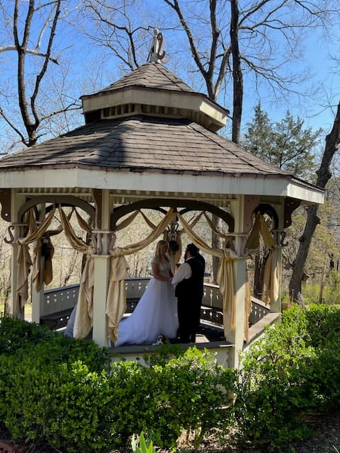 A bride and groom exchange vows inside a decorated gazebo surrounded by trees.
