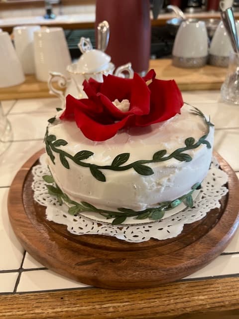 A beautifully decorated cake with white frosting, green leaves, and a red rose on top, placed on a wooden board.