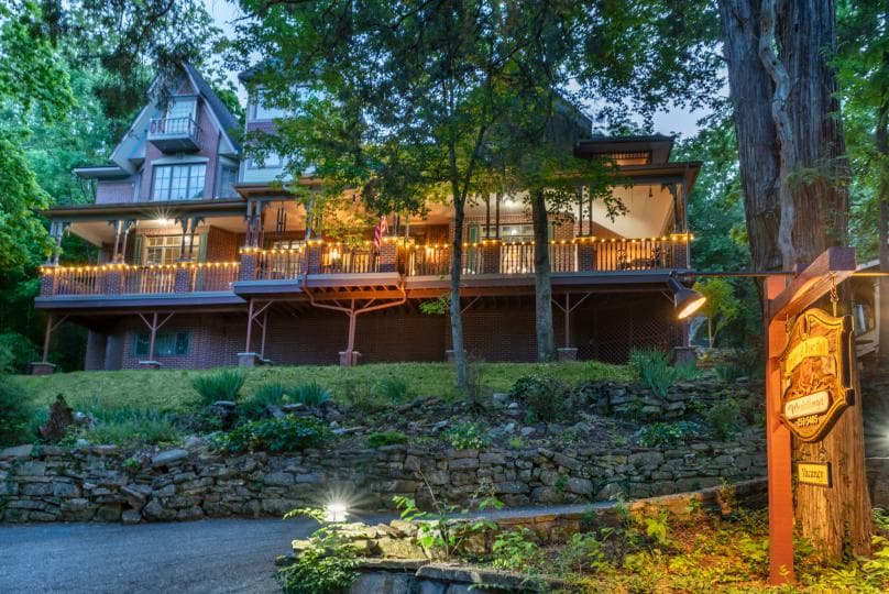 A charming multi-story house with a wooden porch, set amidst lush greenery and illuminated by warm lights.