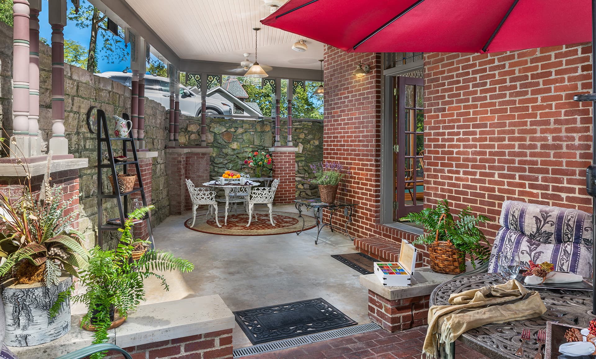 A cozy brick patio with ornate furniture, plants, and a bright red umbrella.
