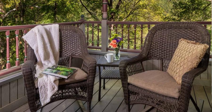 A cozy balcony with two wicker chairs, a small table, a coffee cup, a book, and a vase of flowers, surrounded by greenery.