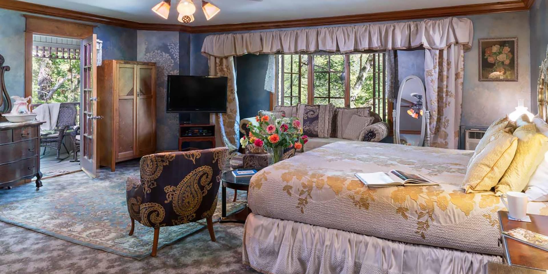 A cozy, elegantly decorated bedroom featuring a queen bed, vintage furniture, and large windows overlooking greenery.