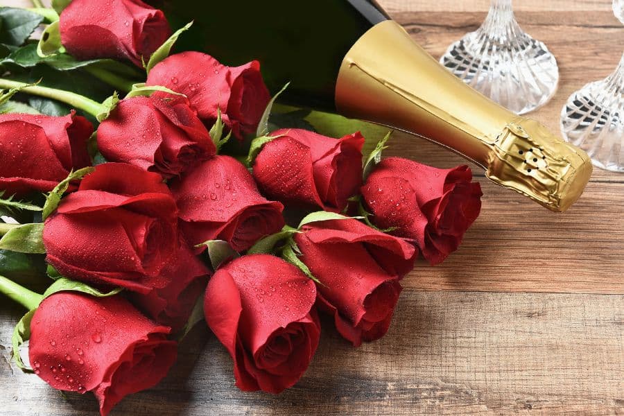 A bouquet of red roses next to a bottle of champagne and crystal glasses on a wooden surface.