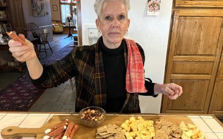 A woman in a plaid jacket holds a snack while surrounded by an assortment of cheeses, crackers, and nuts on a kitchen counter.