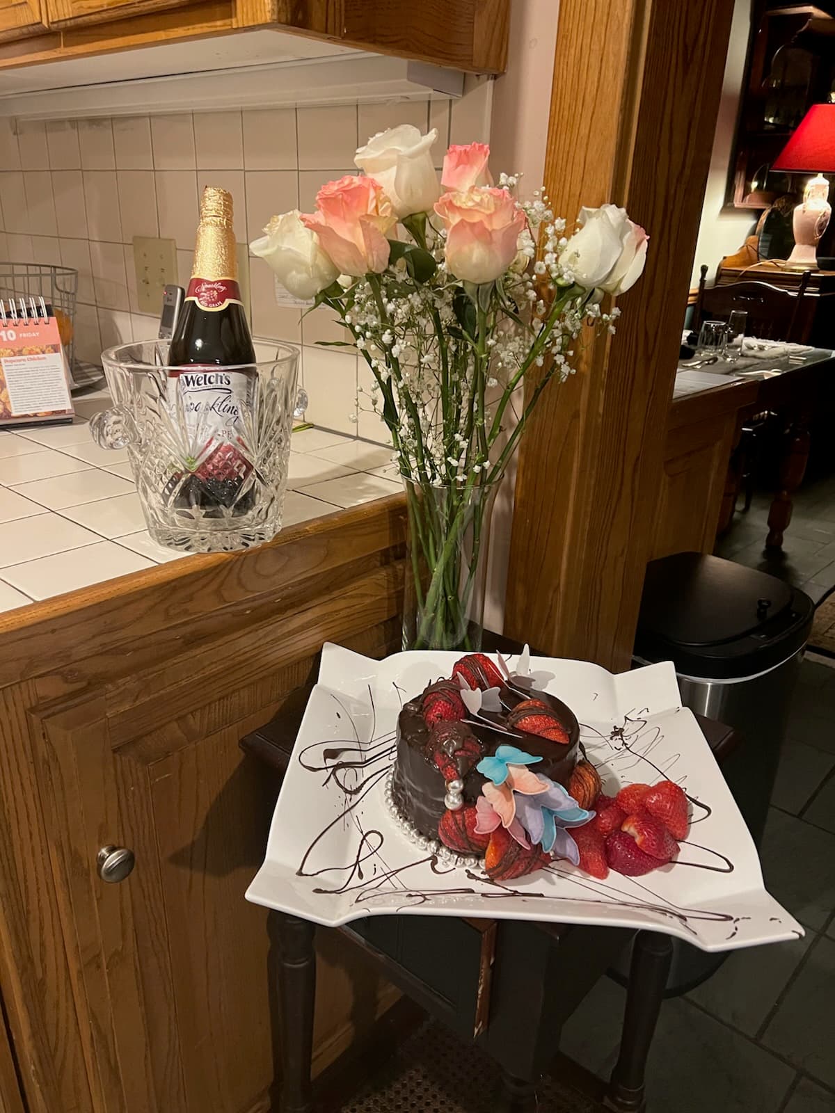 A bouquet of pink roses and baby's breath beside a chilled bottle of sparkling drink and a decorated chocolate cake with fruit on a stylish platter.