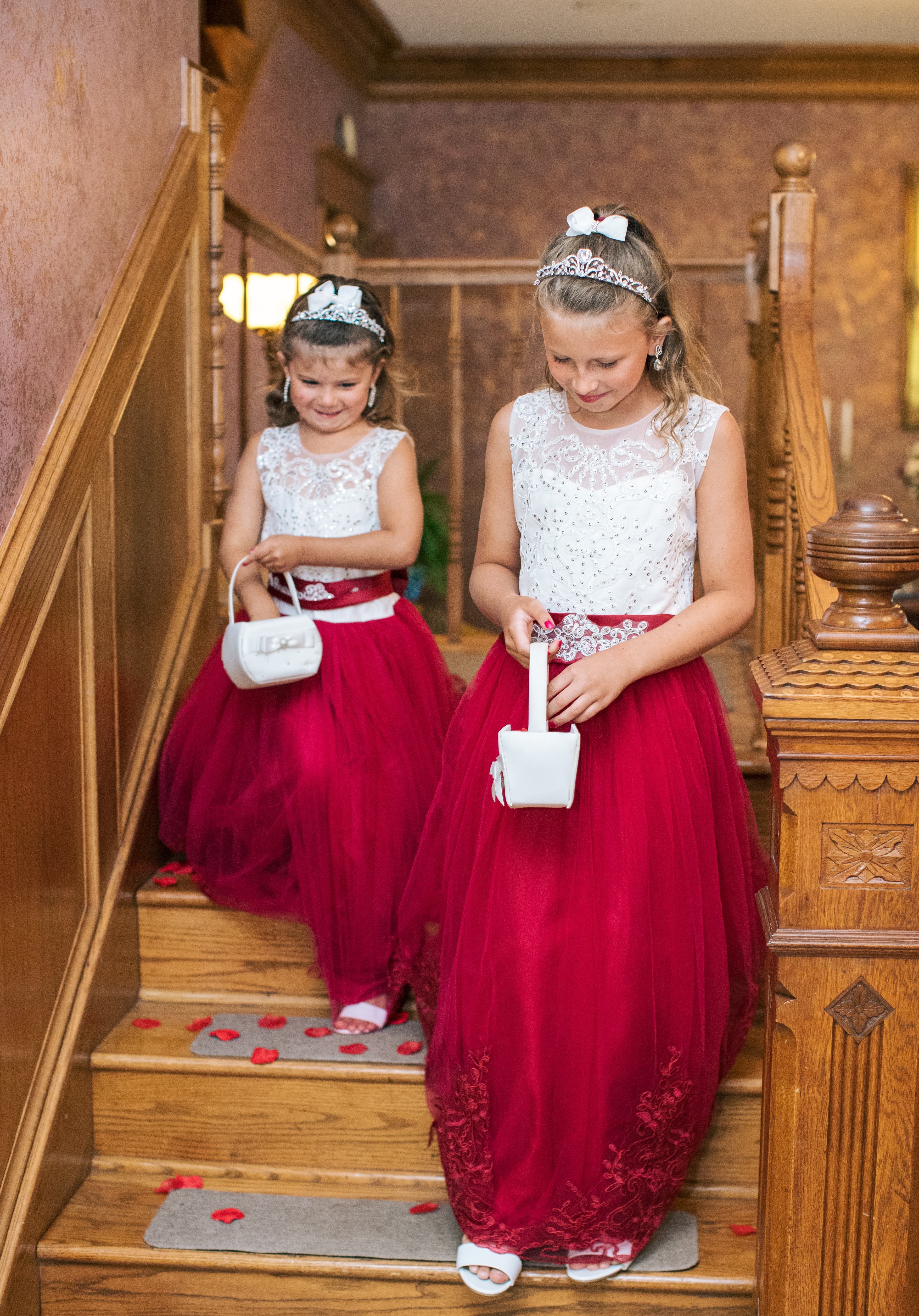 Two young girls in elegant red dresses descend a staircase, each holding a small handbag.