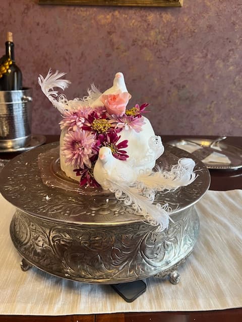 A decorative cake adorned with pink flowers and white bird figurines, displayed on an ornate silver platter.