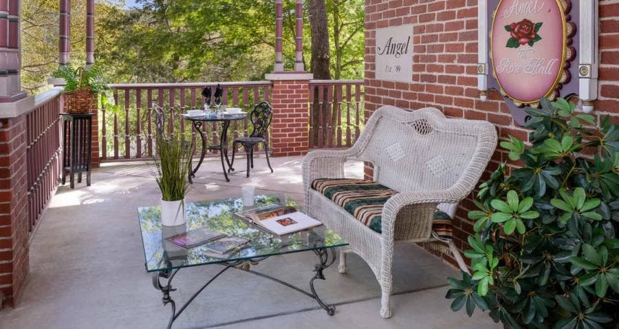 A cozy porch featuring a white wicker sofa, a glass coffee table, and lush greenery.